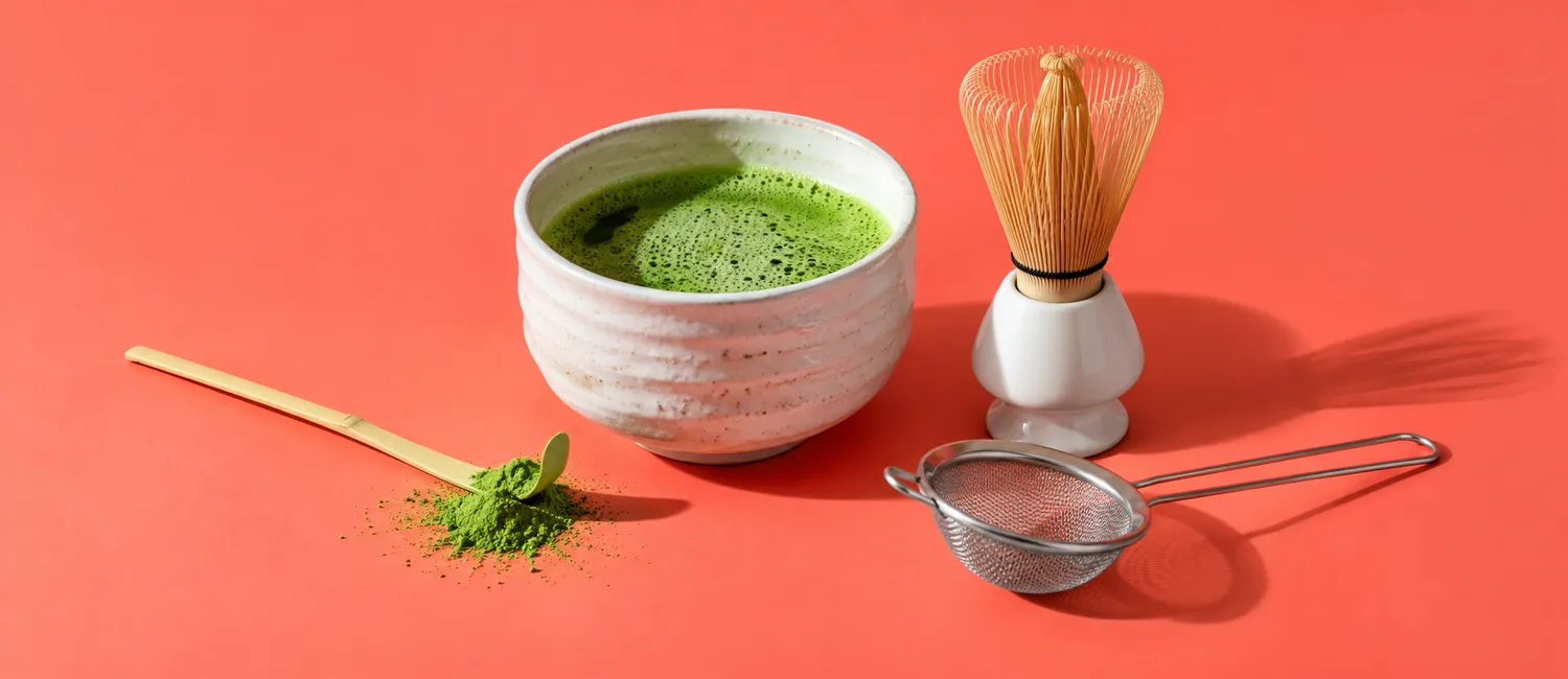Matcha set med bambuvisp, vit keramikskål, bambusked och grönt matchapulver mot korallröd bakgrund