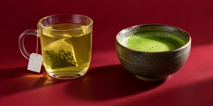 Kopp med grönt te i påse bredvid en skål med matcha te mot vinröd bakgrund