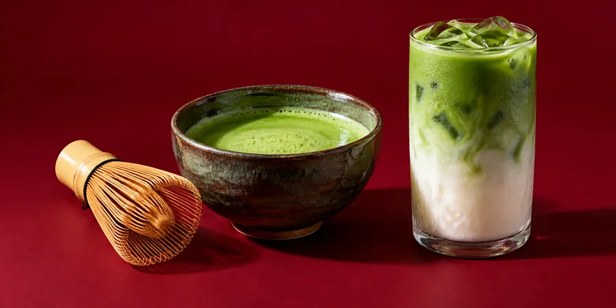 Skål med vispad matcha, bambuvisp och ett glas matcha latte mot vinröd bakgrund